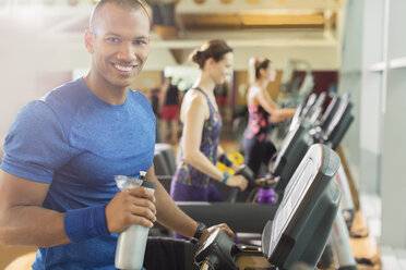 Portrait lächelnder Mann mit Wasserflasche auf dem Laufband im Fitnessstudio - CAIF11822