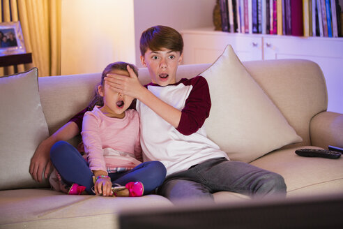 Bruder bedeckt die Augen der überraschten Schwester beim Fernsehen im Wohnzimmer - CAIF11901
