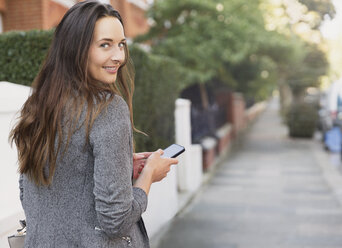 Porträt einer lächelnden Geschäftsfrau mit Mobiltelefon, die auf dem Gehweg zurückblickt - CAIF11907