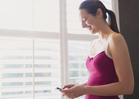 Lächelnde Frau, die mit ihrem Handy am Fenster SMS schreibt, lizenzfreies Stockfoto