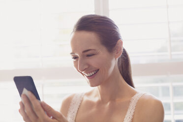 Lächelnde Frau, die mit ihrem Handy am Fenster SMS schreibt - CAIF11910