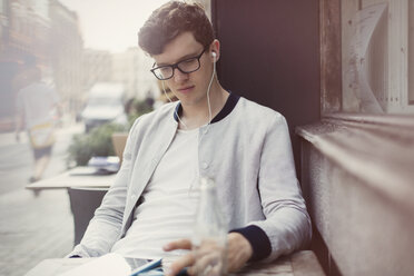 Junger Mann mit Brille und Kopfhörern, der in einem Straßencafé ein digitales Tablet benutzt - CAIF12089
