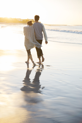 Junges Paar hält sich an den Händen und geht bei Sonnenuntergang am Strand spazieren, lizenzfreies Stockfoto