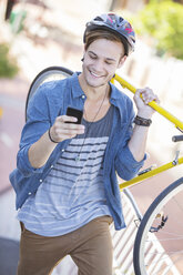 Junger Mann mit Helm, der ein Fahrrad trägt und eine SMS auf seinem Handy schreibt - CAIF12180