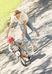 Mutter schiebt ihren Sohn mit Helm auf dem Fahrrad in einem sonnigen Park - CAIF12189