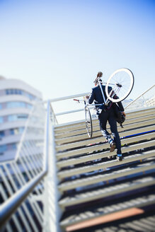 Geschäftsmann, der ein Fahrrad die Treppe hinauf trägt, unter sonnigem blauem Himmel - CAIF12194