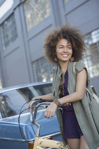 Lächelnde junge Frau mit Afro, die ein Fahrrad auf einer städtischen Straße hält, lizenzfreies Stockfoto