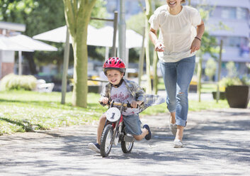 Mutter verfolgt ihren Sohn auf dem Fahrrad mit Helm in einem sonnigen Park - CAIF12212