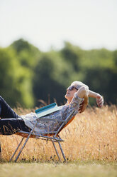 Sorglose ältere Frau entspannt sich mit einem Buch in einem sonnigen Feld - CAIF12218