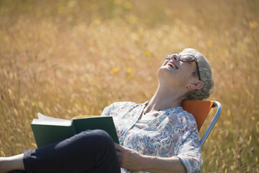 Ältere Frau liest ein Buch und lacht mit zurückgelegtem Kopf in einem sonnigen Feld - CAIF12233