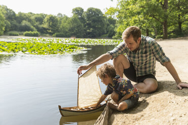 Vater und Sohn spielen mit Spielzeug-Segelboot am Seeufer - CAIF12276