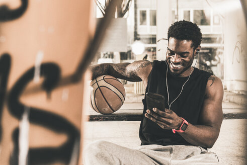 Lächelnder Mann mit Tattoos und Basketball mit Smartphone und Kopfhörern - UUF12978