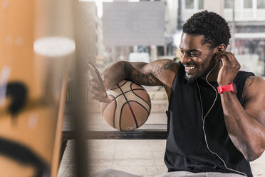 Lächelnder Mann mit Tattoos und Basketball mit Smartphone und Kopfhörern - UUF12979