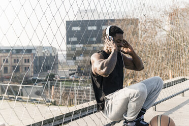 Lächelnder Basketballspieler auf dem Platz, der am Zaun hockt und Musik hört - UUF13017