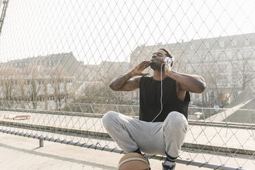 Basketballspieler auf dem Platz, der am Zaun hockt und Musik hört - UUF13018