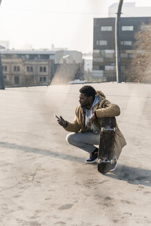 Junger Mann mit Skateboard auf dem Platz, der sein Smartphone überprüft - UUF13027