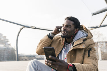 Lächelnder junger Mann mit Smartphone und Kopfhörern im Freien - UUF13031