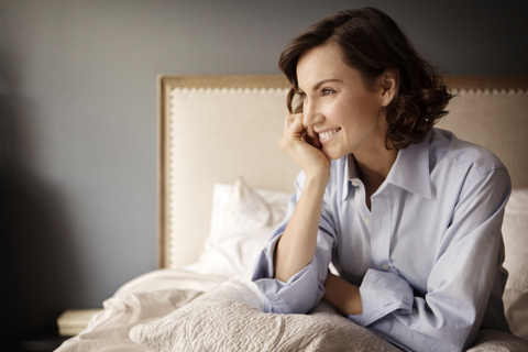 Frau mit Hand am Kinn, die wegschaut, während sie zu Hause auf dem Bett sitzt, lizenzfreies Stockfoto