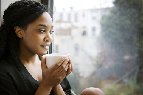 Lächelnde Frau mit Tasse schaut weg, während sie am Fenster sitzt, lizenzfreies Stockfoto