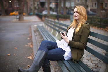 Woman with mobile phone looking away while sitting on bench - CAVF05677