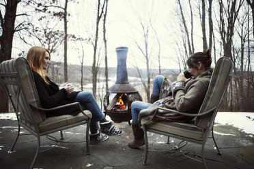 Friends talking and enjoying drink while sitting by bonfire - CAVF05714