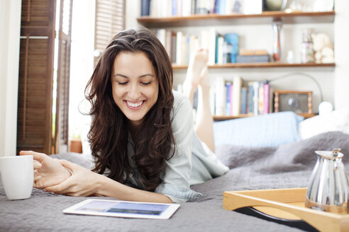 Glückliche Frau schaut auf einen Tablet-Computer, während sie auf dem Bett liegt - CAVF05770