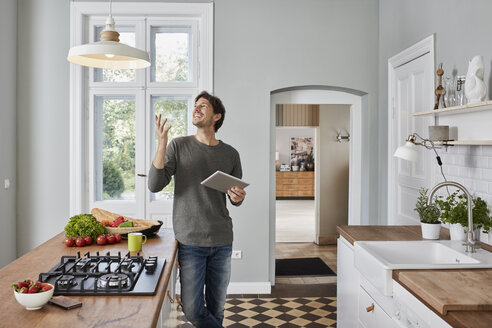 Happy man using tablet in kitchen looking at ceiling lamp - RORF01129