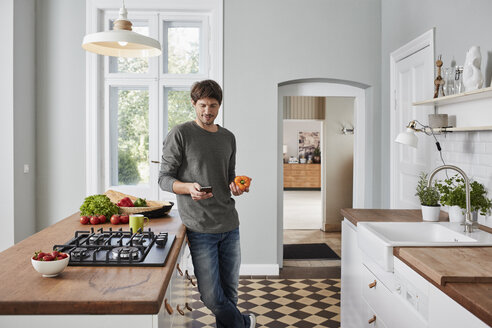 Man using smartphone and holding bell pepper in kitchen - RORF01138