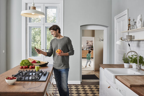 Smiling man using smartphone and holding bell pepper in kitchen - RORF01139