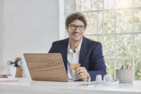 Porträt eines lächelnden Geschäftsmannes mit Laptop auf dem Schreibtisch, der eine Kreditkarte hält - RORF01148