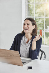 Lachende Geschäftsfrau mit Laptop auf dem Schreibtisch, die eine Karte hält - RORF01154