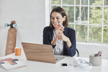 Happy businesswoman using laptop on desk holding card - RORF01157