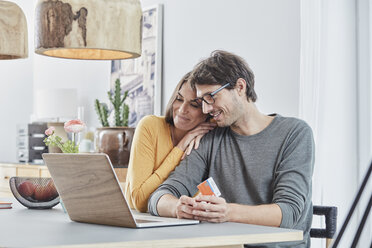 Smiling couple with a card using laptop on table at home - RORF01165