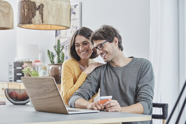 Happy couple with a card using laptop on table at home - RORF01166