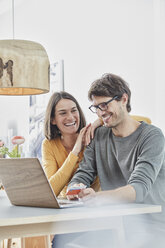 Happy couple with a card using laptop on table at home - RORF01167