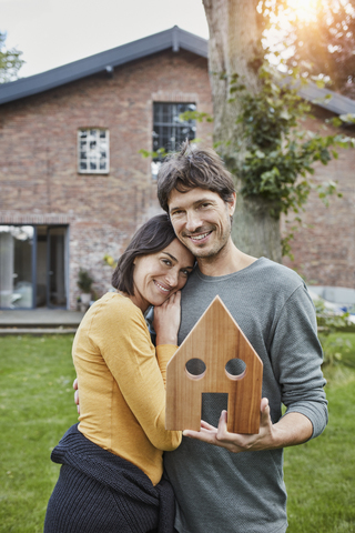 Porträt eines lächelnden Paares im Garten ihres Hauses mit Hausmodell, lizenzfreies Stockfoto