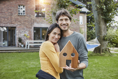 Porträt eines lächelnden Paares im Garten ihres Hauses mit Hausmodell, lizenzfreies Stockfoto