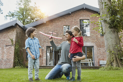Vater mit zwei Kindern, die mit einem Spielzeugflugzeug im Garten ihres Hauses spielen - RORF01207