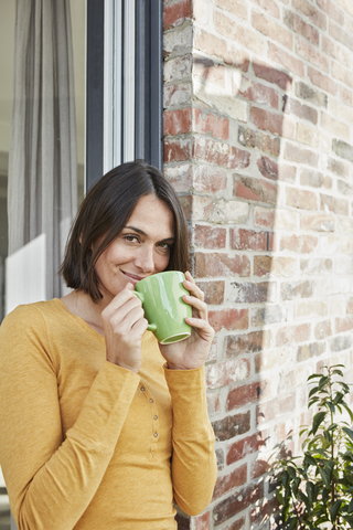 Porträt einer lächelnden Frau, die vor ihrem Haus aus einer Tasse trinkt, lizenzfreies Stockfoto