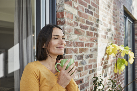 Lächelnde Frau, die vor ihrem Haus aus einer Tasse trinkt, lizenzfreies Stockfoto