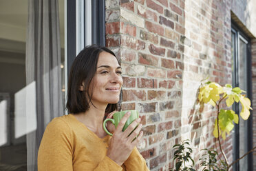 Smiling woman drinking from cup in front of her home - RORF01214