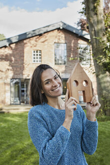 Portrait of smiling woman in garden of her home holding house model - RORF01237