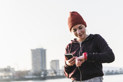 Sportliche junge Frau am Flussufer in der Stadt mit Smartphone und Smartwatch, lizenzfreies Stockfoto