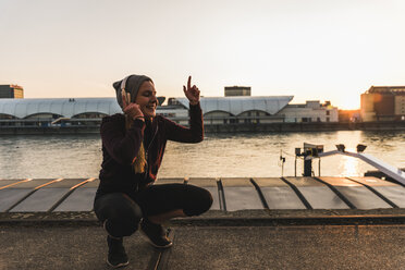 Fröhliche sportliche junge Frau, die am Flussufer in der Stadt bei Sonnenuntergang Musik hört - UUF13083