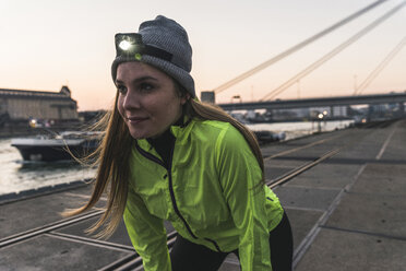 Sportive young woman with headlamp at dusk at the riverside in the city - UUF13084