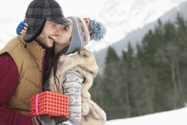 Glückliches Paar mit Weihnachtsgeschenk, das sich im verschneiten Feld küsst - CAIF12360