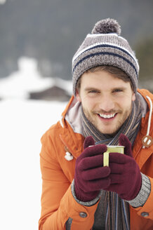 Nahaufnahme eines lächelnden Mannes mit Strickmütze und Handschuhen, der in einem verschneiten Feld Kaffee trinkt - CAIF12368