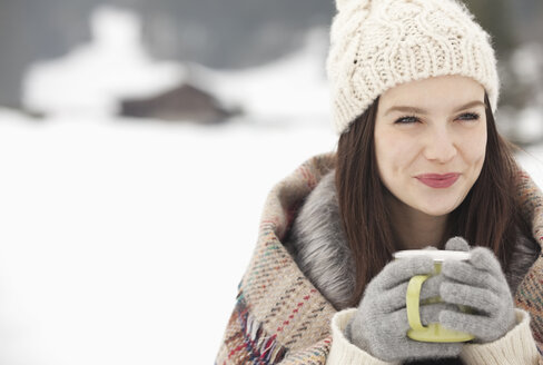 Nahaufnahme einer lächelnden Frau mit Strickmütze und Handschuhen, die in einem verschneiten Feld Kaffee trinkt - CAIF12369