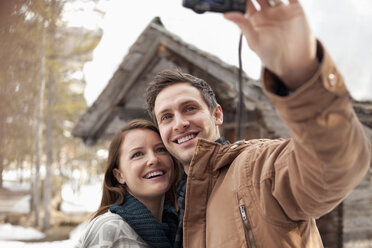 Smiling couple taking self-portrait with digital camera outside snowy cabin - CAIF12399