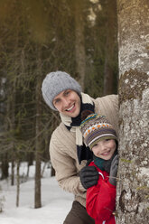 Porträt von glücklichem Vater und Sohn hinter einem Baumstamm im verschneiten Wald - CAIF12416